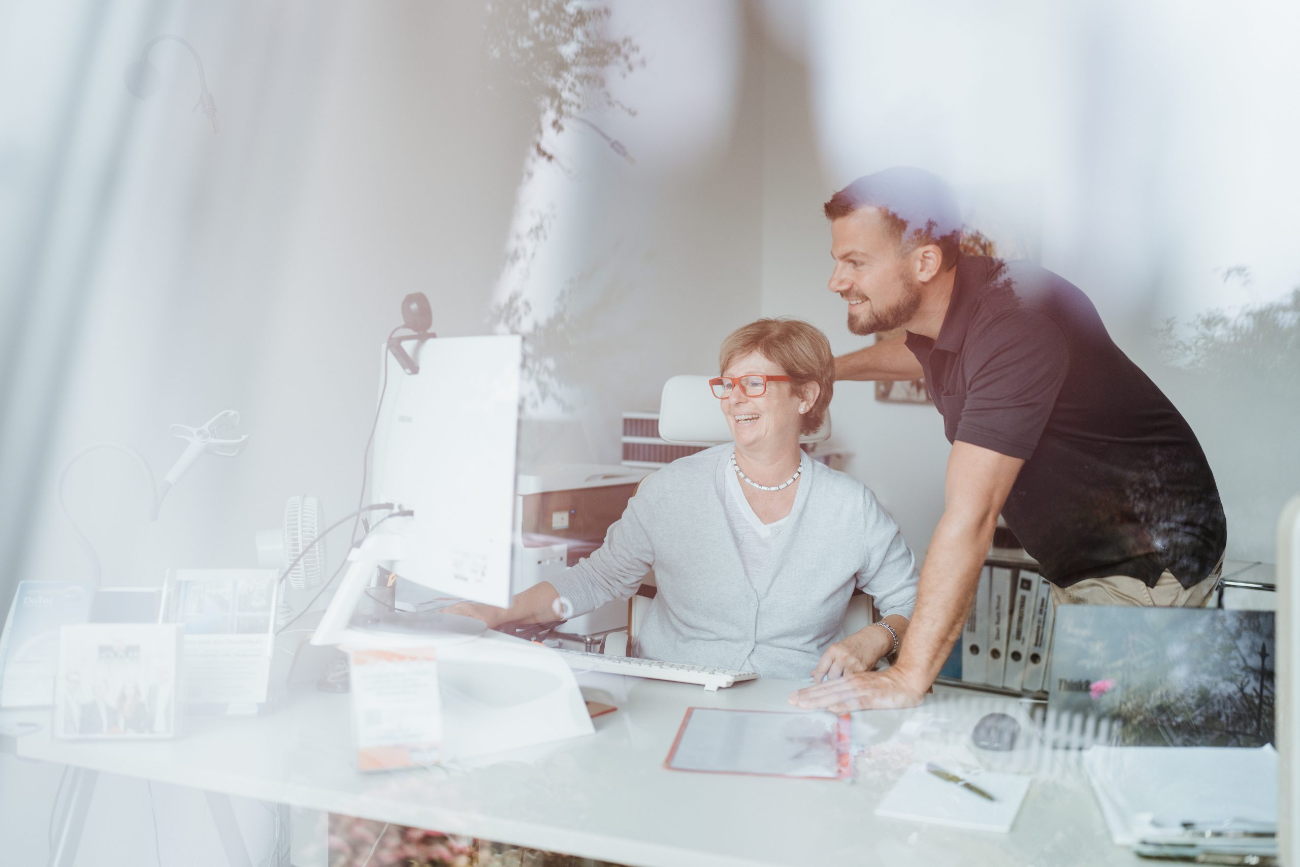 Frau mit Brille bedient einen Desktop-Computer, ein Mann lehnt sich über den Schreibtisch und blickt auf den Monitor in einer Büroumgebung. 