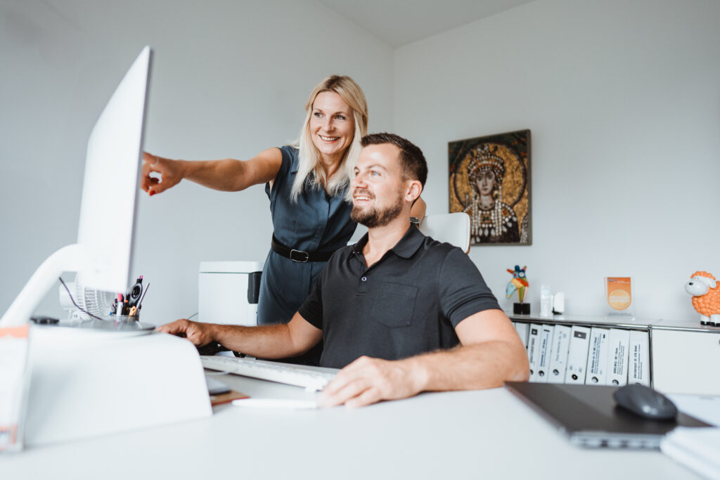 Frau zeigt auf Monitor, während ein Mann am Schreibtisch in einem hellen Büro am Computer arbeitet 