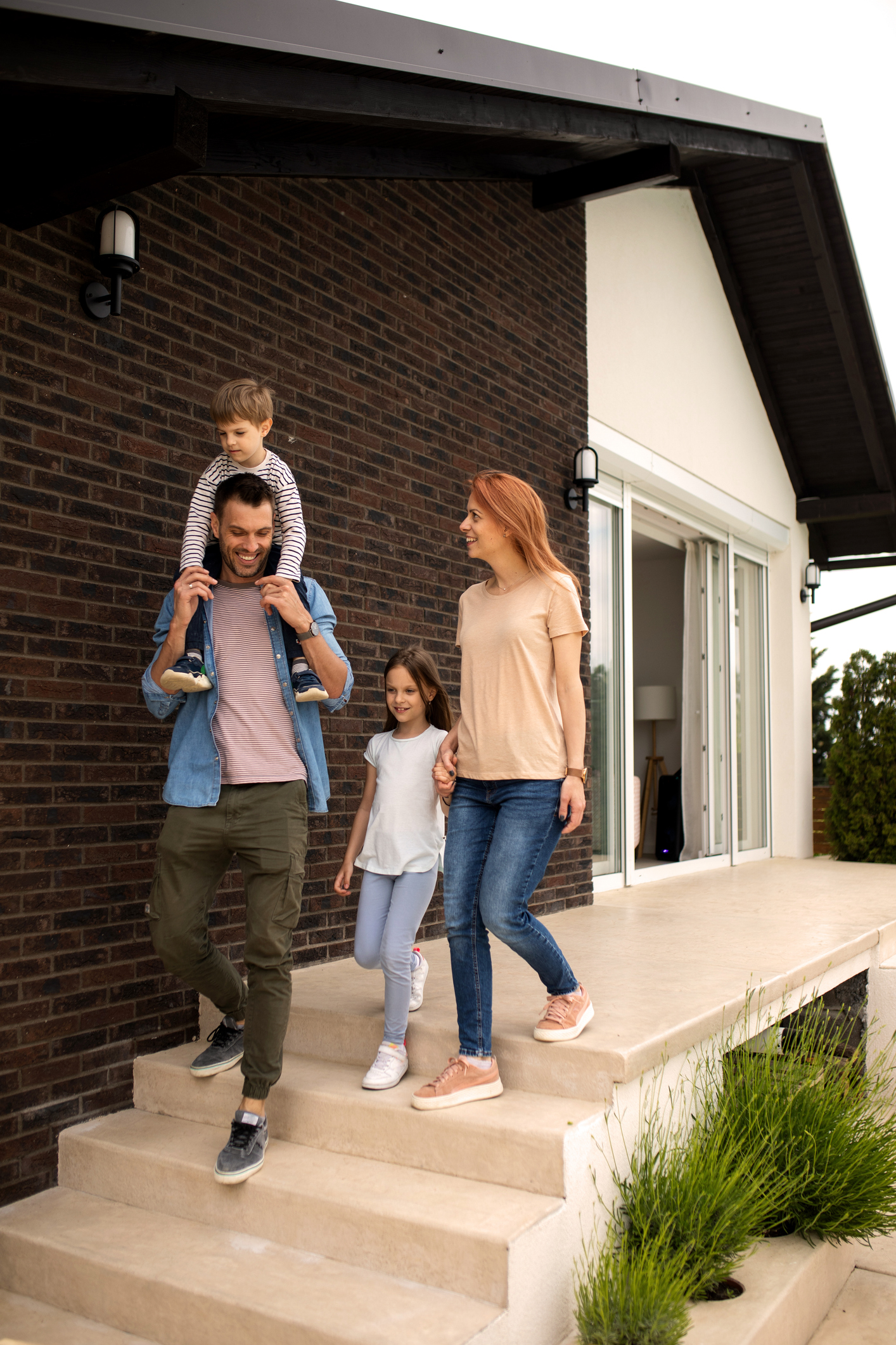 Familie mit zwei Erwachsenen und zwei Kindern geht gemeinsam eine Treppe vor einem modernen Einfamilienhaus hinunter. 